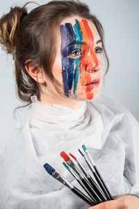 Young woman with painted face holding paintbrushes against gray background