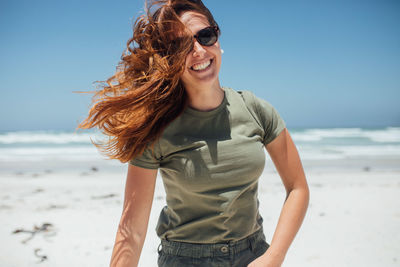 Cheerful young woman wearing sunglasses while standing at beach during sunny day