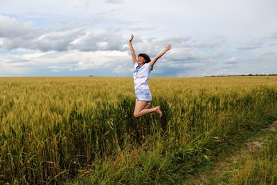 Cheerful young woman with arms raised jumping on field against cloudy sky