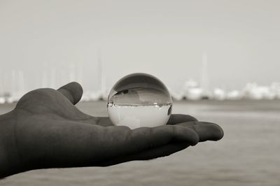 Close-up of hand holding crystal ball by sea against sky