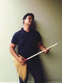 Young man looking away while holding broom against wall