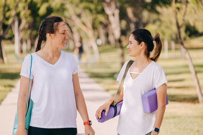 Two smiling young women walking in the morning park. carrying mats and other equipment for yoga.