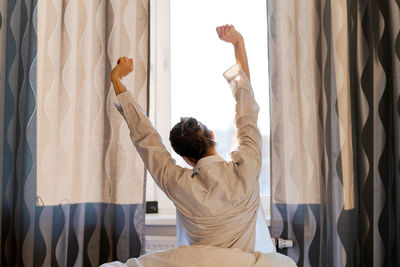 Back view caucasian woman stretching arms and body near window after waking up