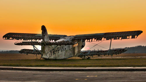 Damaged and abandoned airplane on field against orange sky during sunset