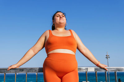 Concentrated ethnic female athlete with curvy body and closed eyes in active wear against fence and ocean in sunlight