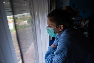 Close-up of woman wearing mask looking through window at home