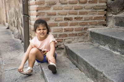 Girl making face while sitting on footpath against brick wall