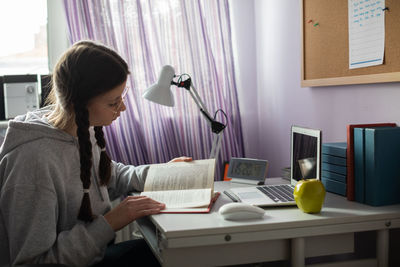 A student with braids and glasses at home reads a book.
