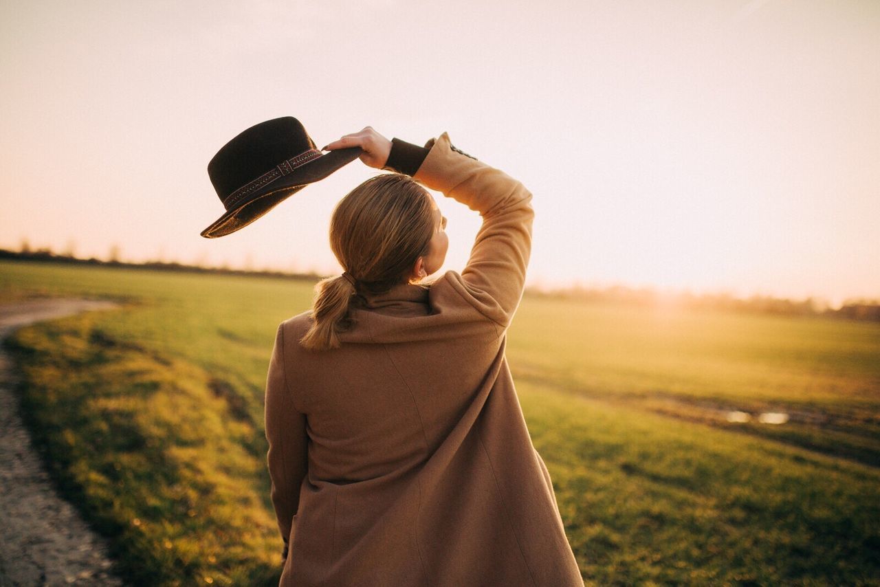 Rear view of woman holding hat on grassy field against sky