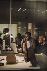 Smiling male and female colleagues discussing over laptop while working last minute in office at night