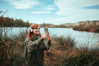 Young woman taking selfie with smart phone while standing on land against lake