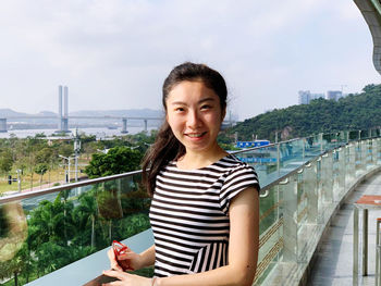 Portrait of smiling young woman standing by railing against sky