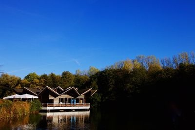 House and trees by lake against blue sky