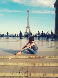 Side view of woman sitting by steps while looking at eiffel tower against sky