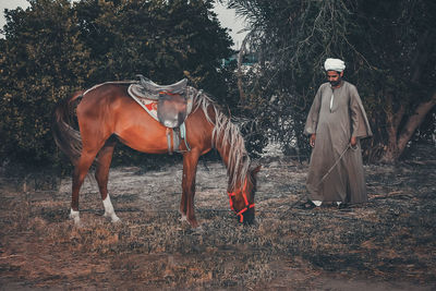 Full length of man in traditional clothing standing by horse grazing on field