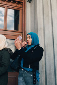 Low angle view of young muslim woman applying lipstick looking into mobile phone by friend against building