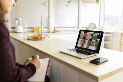 Young working woman with notebook doing video call with colleagues on laptop at home