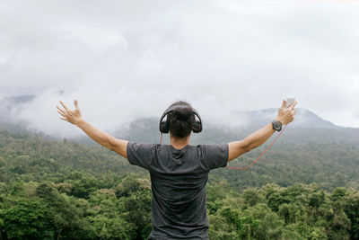 Rear view of man with arms raised listening music on mobile phone while standing against mountain and sky