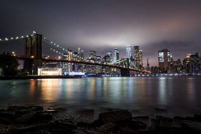 Illuminated bridge over river by buildings against sky at night