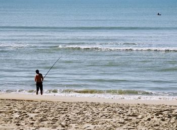 Rear view of shirtless man fishing in sea while standing on shore at beach