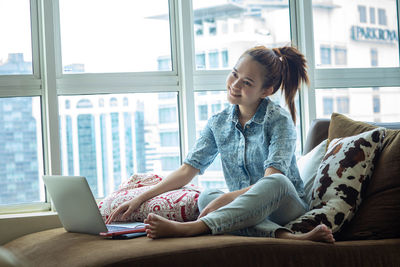 Young woman using phone while sitting on window