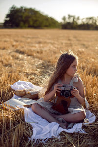 Girl child in a dress sitting on a mown field with a camera on a blanket with bread and a book