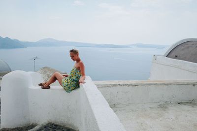 Young woman sitting on retaining wall by sea against sky