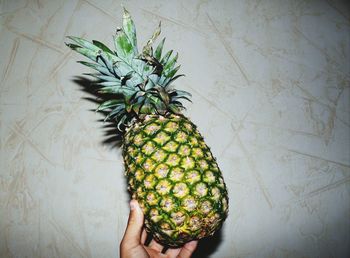 Cropped hand of person holding pineapple against wall