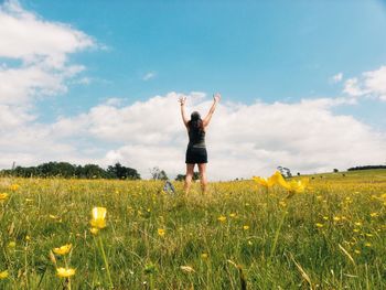 Rear view of woman with arms raised standing amidst yellow flowers on field