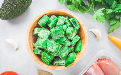 Frozen diced spinach in a wooden bowl with avocado and fresh parsley on a light gray cement.