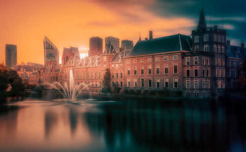 Illuminated buildings by river against sky during sunset