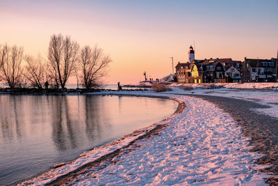 Bare trees by river against sky during sunset