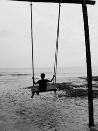 Rear view of boy enjoying swing at beach against sky