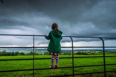 Rear view of girl standing on fence by grassy field against cloudy sky