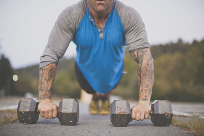 Low section of man with dumbbells doing push-ups on road at park