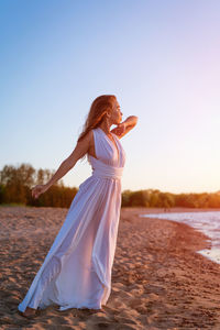 Young woman in summer in a white dress on the shore at sunset