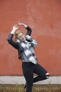 Portrait of happy woman standing on one leg with arms raised against brown wall