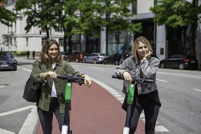 Portrait of smiling female friends with electric push scooters standing on street in city