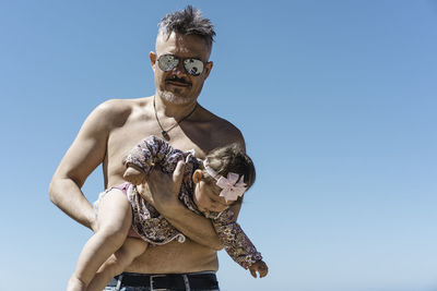 Midsection of shirtless man holding sunglasses against clear sky