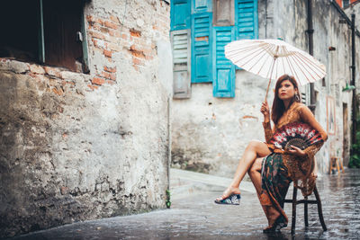 Full length of beautiful woman with umbrella sitting on stool in old town