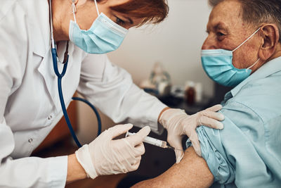 Doctor holding syringe with vaccine and making injection to senior patient with medical mask