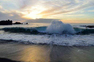 Waves splashing on shore at beach against sky during sunset