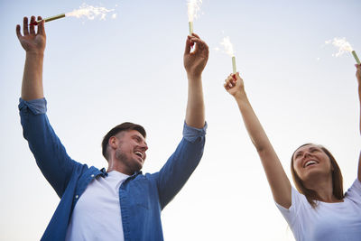 Low angle view of smiling couple holding lit sparklers against clear sky