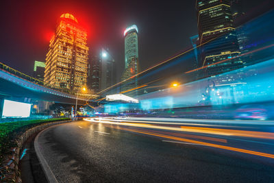 Light trails on road by illuminated buildings against sky at night