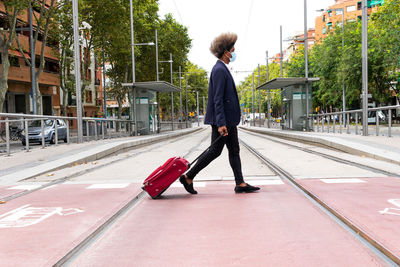 Side view of african man with afro hair and a mask walking with a suitcase on wheels while crossing a tram track in a city