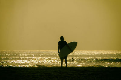 Rear view of silhouette man with surfboard standing on shore at beach during sunset