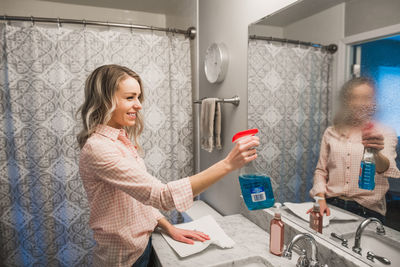 Side view of smiling young woman cleaning mirror in bathroom