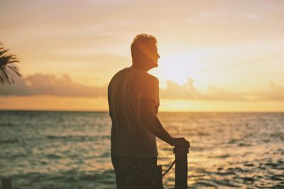 Rear view of young man looking at sea while standing against sky during sunset