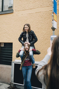 Female photographing girl carrying friend on shoulder against wall in city