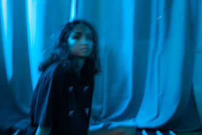 Young woman looking away while standing against blue curtain